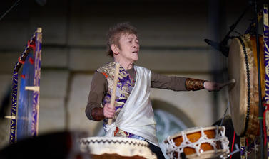 Figure playing the drums, one arm outstretched to his left