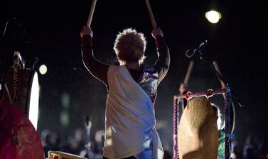 Figure from behind playing the drums, arms raised above his head with drumsticks in his hands