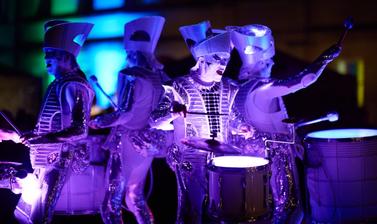 Figures elaborately dressed and covered in lights play the drums