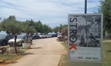 Image depicts the Istrox Exhibition Poster in from to Lapidarium building nex to a small road and cars parked in the background flanked by trees.