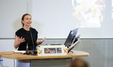 Someone stood behind a podium, mid speech and gesturing with both hands splayed