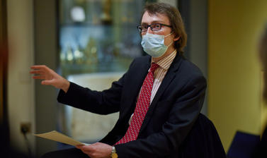 Someone in a suit sat down, gesturing with one hand and holding paper in the other