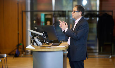 Someone stood behind a podium, pointing upwards with both hands.