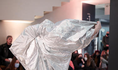 A photo of a Butoh dancer, holding a silver metallic sheet wide behind her back.