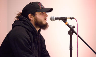 Performer in a black hoodie and hat leaning towards a microphone