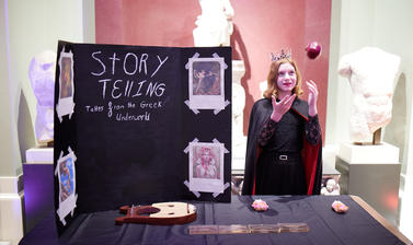 A woman dressed as a queen throwing an apple. They are stood behind a table with a sign reading 'story telling'
