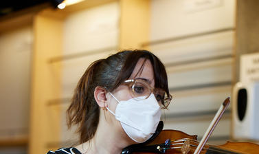 Brunette, female musician, sat down, wearing black top with white pattern, playing violin.
