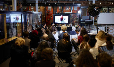 Image of a panel interview, including Zoom call speaker, with life audience and based at a museum hall.