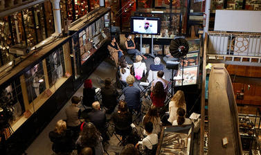 Birdseye view of  a panel interview, including Zoom call speaker, with life audience and based at a museum hall, surrounded by objects displayed by the museum. . 