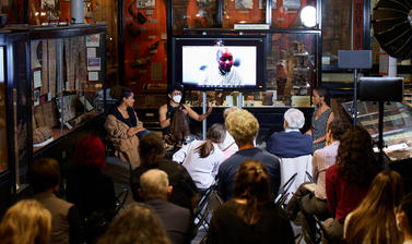 Image of a panel interview, including Zoom call speaker, with life audience and based at a museum hall. Three panellists and Zoom call speaker, are all seen facing the camera and life audience from the back.