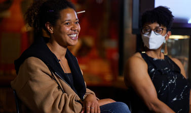Two female panellists, captured whilst sat down, with blurred museum objects in the background. Both females are brunette, one with curled pony tail and smiling, one with short hair and wearing white face mask.