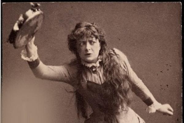 Monochrome image depicts Adeleide Johannessen in character as Nora dancing the tarantella dance and holding, above her head, a tambourine in her right hand.