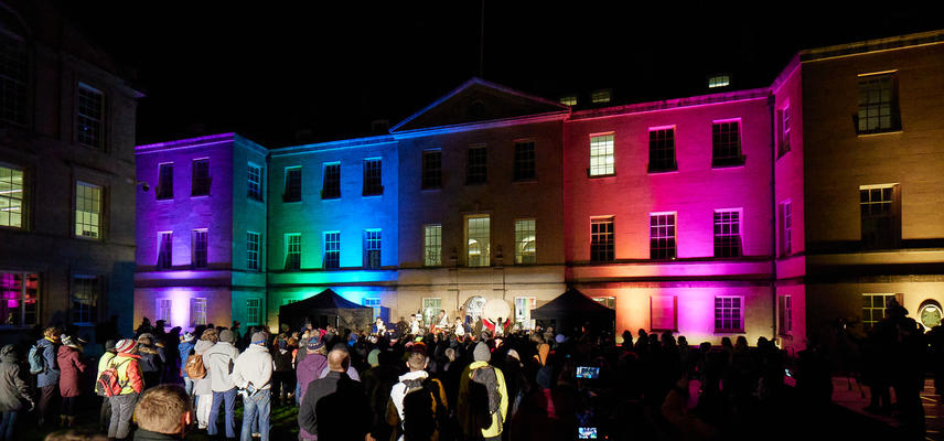 A crowd watches drummers, with the building behind the drummers lit up in different colours.