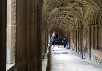 Stone cloisters - stone columns rising to intricate stone ceiling. View down a long cloister hallway with a family in the distance; three figures walking, with one adult carrying a child on their shoulders. 