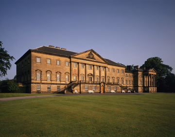 Exterior shot of Nostell Priory. A large building of warm-coloured stone, imitating Classical architecture with columns and pediment, and 21 sets of triple windows. The foreground of the image is a green lawn. 