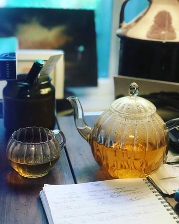 Glass teapot and cup with tea sitting on a table with open notebook in front 