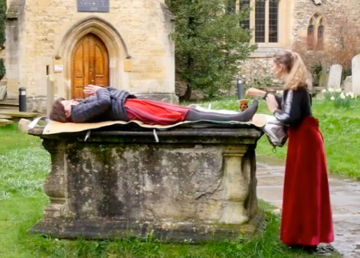 Man lies on ancient tomb. Woman in red skirt stands beside him.