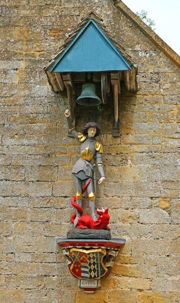 The statue of St George with the dragon between his feet stands on a corbel that bears a coat of arms. St George has his right hand risen close to the bell, which hangs over his head. A small wooden roof protects both the bell and the statue from rain. 