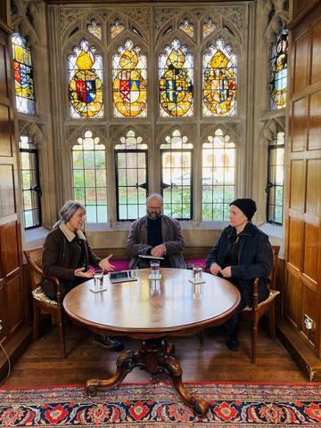 Three people sit around a table in front of a large stain glass window. The man sits in the centre with Alice Oswald and Katie Mitchell on either side.