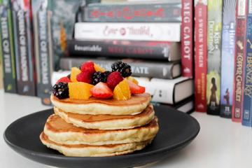 Staple of pankakes topped with berries arranged on a plate standing in front of a row of books.