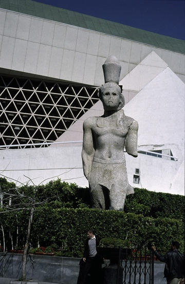 Frontal photograph of a colossal Egyptian statue in grey stone. In the background the architecture of the library, with various triangles introduced in the design, is evokes the pyramids. 