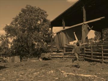 Sepia still from digital video 'Stip Lander' depicting Samson Kambalu running with streched out arms, imitating a landing aircraft, in a wooden plank fenced field flanked by a large tree and a open spaced barn. 