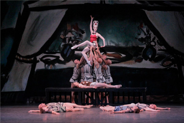 Image depicting four dancer laying on the floor, another one laying on a bench in fron of them and behind are nine dancers standing close togherther in a circle lifting a ballerina in a stitting pose wearing a crown.