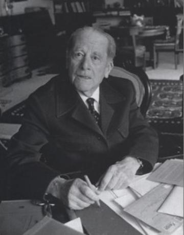 A black and white photo of an old man in a suit at a desk with letters around him