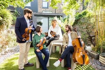 4 musicians with stringed instruments sitting around a garden table talking
