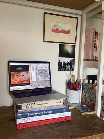 An open laptop balanced on a pile of books relating to the Bloomsbury Group. These items are placed on a desk with a jar of pens and pencils and decorative tiles, with art posters hanging on the wall above.