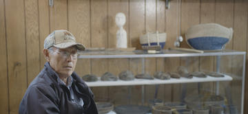 An elderly Yup’ik man giving an interview. In the background are masks and other artefacts at the Nunalleq Museum in Quinhagak.
