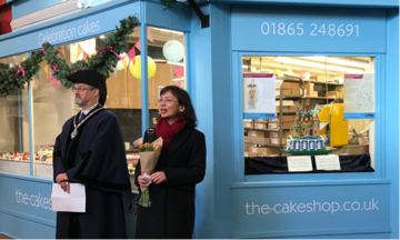 A woman introducing steve goddard, stood in front of a cake shop