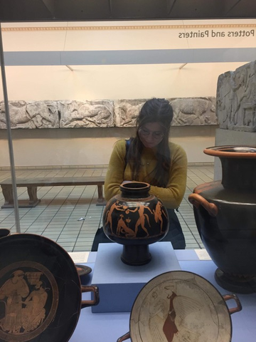 Abi examines a Greek vase in a museum setting. She has her arms crossed and is looking at it thoughtfully. 