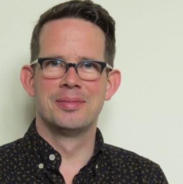 man with brown hair and thick black glasses smiling at camera