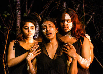 two women of colour hold and look at Medea (middle)who laments. White tribal markings on faces, wearing plain black clothing. Trees are in the background. 
