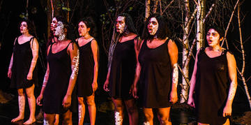 actors of colour from the chorus of medea line up next to each other. All wear black, with tribal markings on their face in white