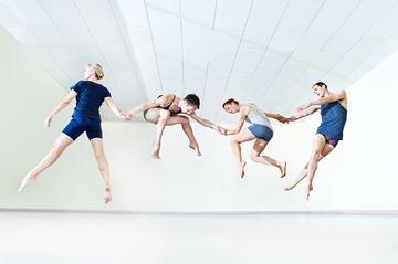 In a dance studio four dancers are in mid-air, holding hands with each dancer in a different pose (straddle, squat, sprint, and stretched legs crossed).