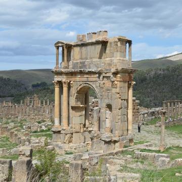 Alt text: View of Roman arch, with remains of the columns of the city plaza to its right, and hills in the background.