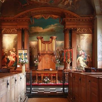 Image depicting nave of St Lawrence, Whitchurch Lane, Little Stanmore - East end with wooden pews, the altar in front of the organ flanked by a set of wooden columns (square and round). The walls depict religious cenes framed with ornamental wood carvings
