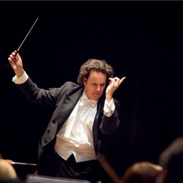 Conductor dressed in formal concert dress with baton in hand and arms raised mid conducting
