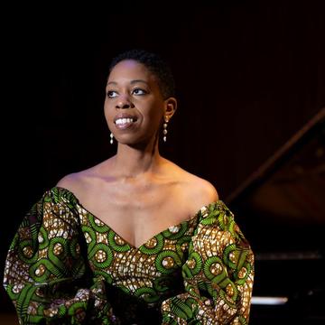 Woman in african patterned dress stares to the left while a piano is in the background to the right