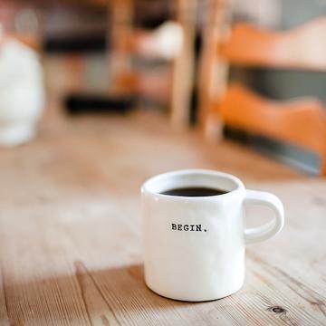 A white mug of coffee sat on a wood table. The mug has 'BEGIN.' printed on it.