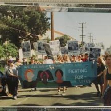 A blue banner with three female faces leads the front of a protest march along a street with people holding signs behind it
