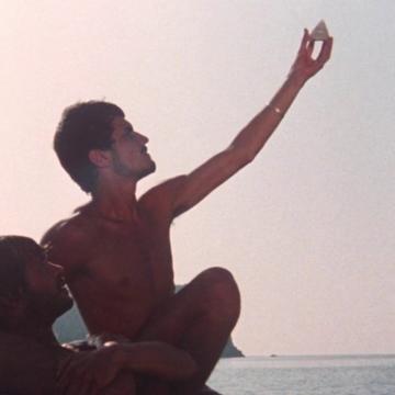One topless man supports another holding a triangle up in the air on a sea-scape background.