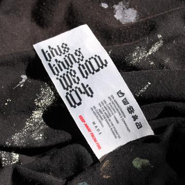 A colour photograph of a clothing tag at the centre, which reads "this thing we call art".  The tag is attached to a wrinkled black t-shirt with white, yellow, green, blue, and orange paint splatter stains on it.