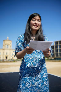 Close up of Jenny Wong wearing a blue dress and reading from a page on a sunny day in front of the Radcliffe Observatory