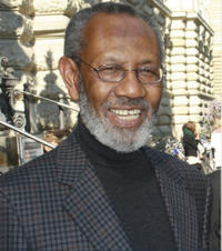 abdilatif abdalla he is smiling at the camera. he has a beard and is wearing a dark roll neck with a dark checked jacket