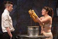 2 people stood facing each other, one is wearing a white shirt and black trousers with her hair tied back. The other is wearing flesh coloured underwear their hands are covered in gold paint lifting from a bucket
