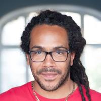 Man looking at the camera with black rimmed glasses, dreadlocks, and a red tshirt