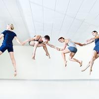 In a dance studio four dancers are in mid-air, holding hands with each dancer in a different pose (straddle, squat, sprint, and stretched legs crossed).